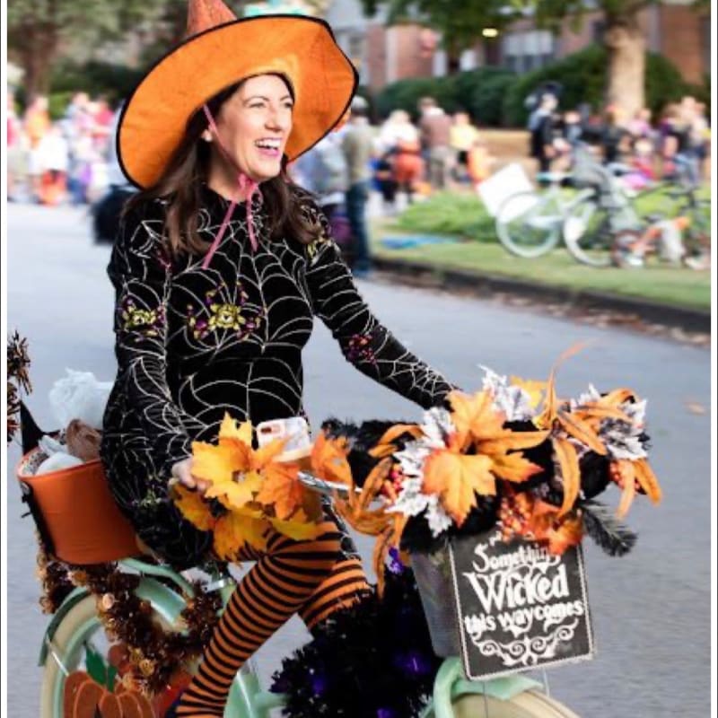 A woman in a witch costume rides a decorated bicycle with autumn leaves and Halloween signs, while a blurred crowd and trees form the background.