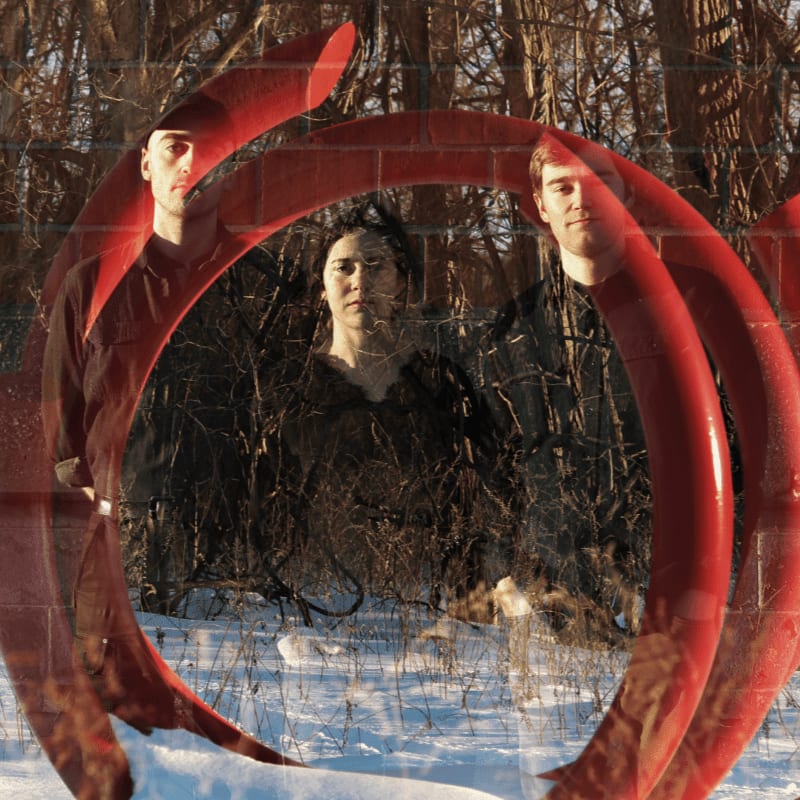 Three people stand in a snowy wooded area, partially obscured by a large, red, abstract sculpture and a brick pattern.