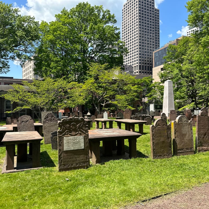 A historic graveyard with weathered headstones and tables in the foreground is set against a backdrop of lush green trees and modern skyscrapers under a cloudy sky.