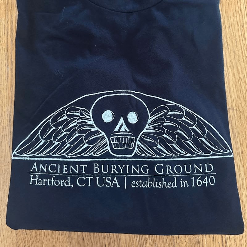 A dark t-shirt with a white winged skull emblem and text reading "ANCIENT BURYING GROUND Hartford, CT USA | established in 1640" lies on a wooden surface.