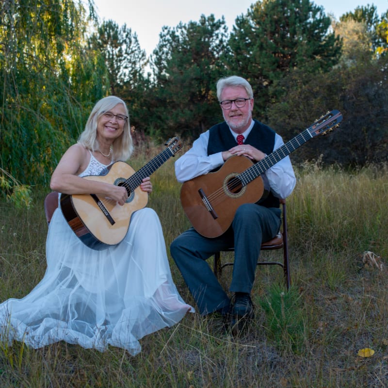 A smiling couple, a woman in a white dress and a man in a vest, sit outdoors with their guitars in the foreground, while a backdrop of trees and foliage frames them.