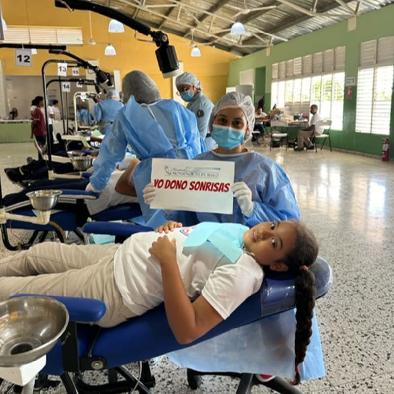 A young girl lies in a dental chair in the foreground, while a masked dentist holds a sign in the background of a clinic.