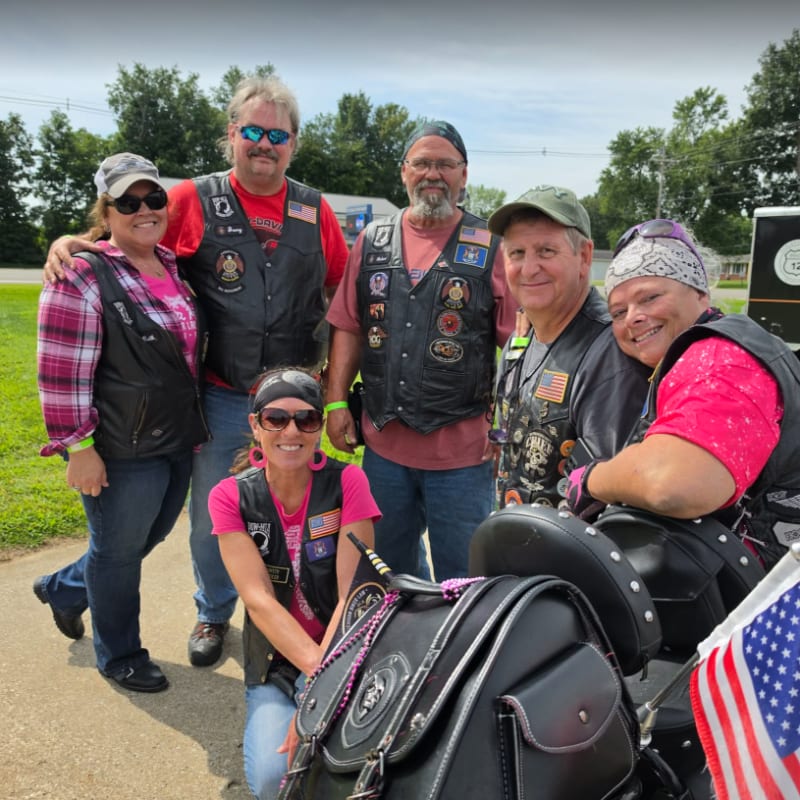 A group of six smiling people, some wearing leather vests and bandanas, pose in front of a motorcycle with an American flag, with a grassy area and trees in the background.