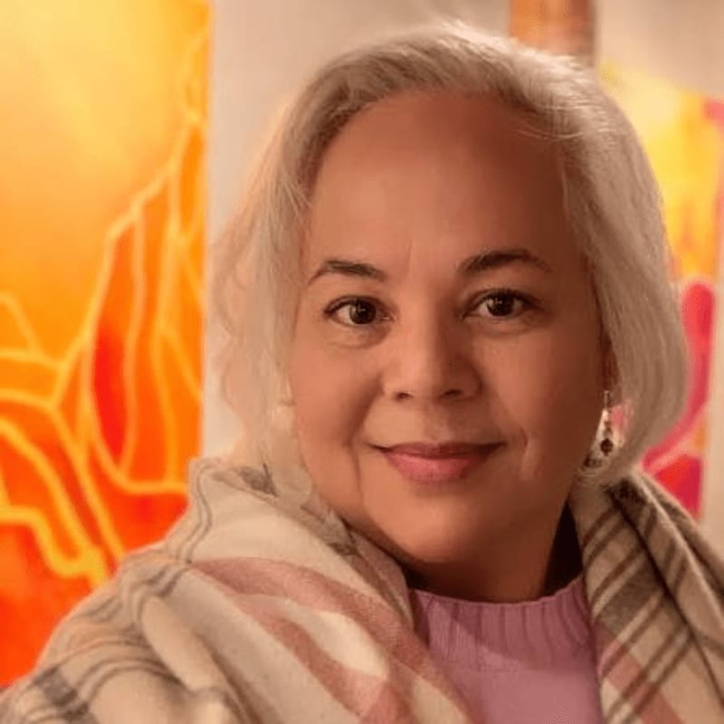 A woman with gray hair and a light purple shirt is wrapped in a patterned scarf, with abstract orange and yellow artwork in the background.