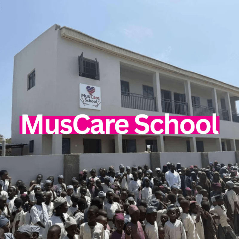 A large crowd of children in traditional attire gathers in front of a light-colored school building with a pink "MusCare School" banner.