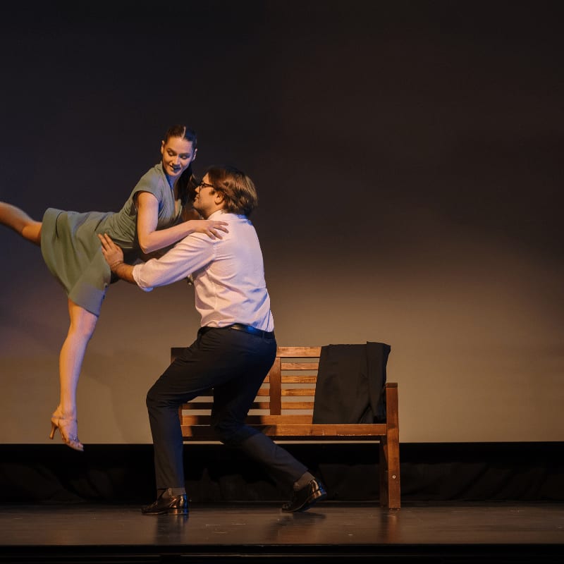 A man in a pink shirt and dark pants supports a woman in a teal dress as she dances, with a wooden bench and a jacket in the background.
