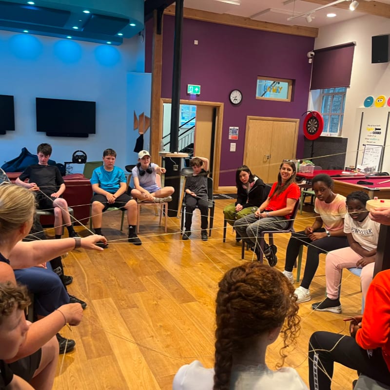 A group of young people sit in a circle on chairs in a brightly lit room, holding onto a rope, with a dartboard and pool table visible in the background.