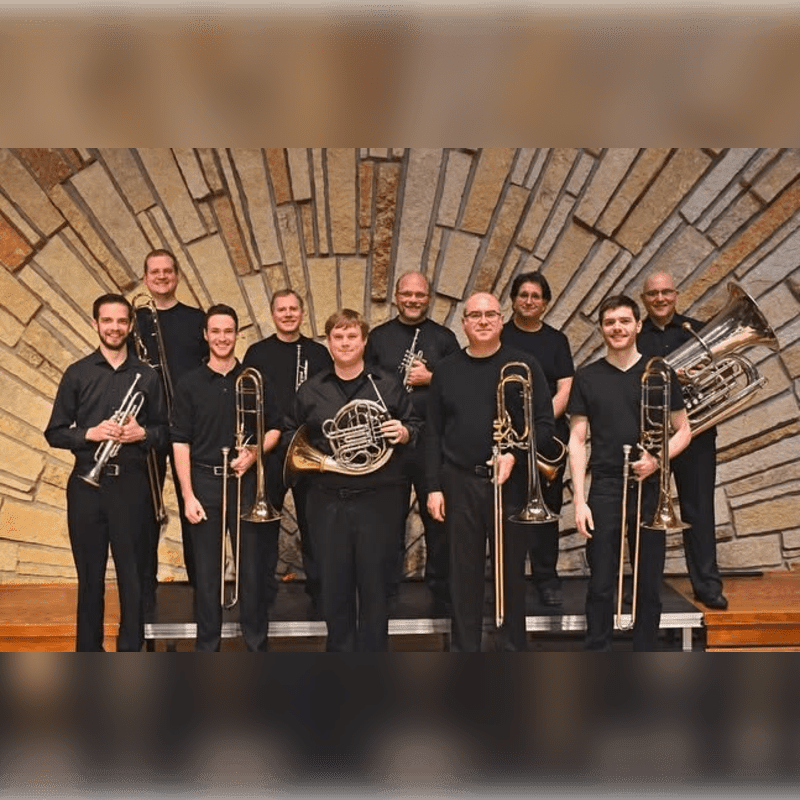 A brass ensemble stands in front of a textured stone wall, with musicians holding various instruments.