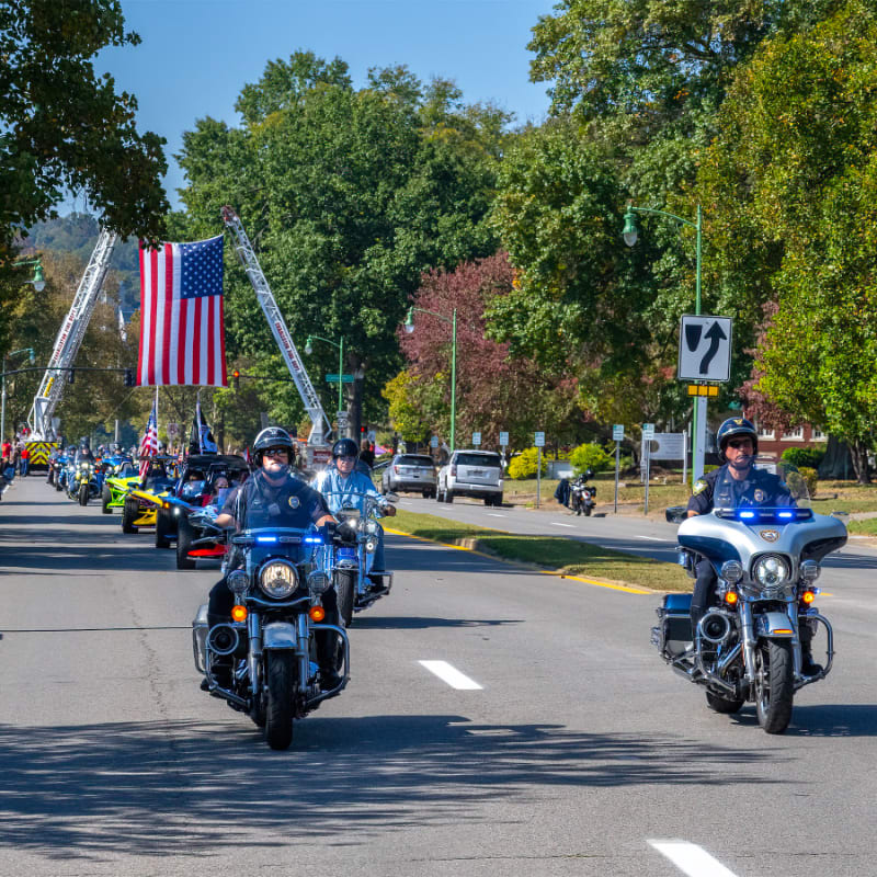 Police officers on motorcycles lead a parade with a fire truck and American flag in the background under a clear blue sky.