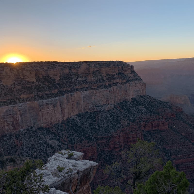 A rocky ledge in the foreground overlooks the vast, layered canyon bathed in the warm glow of a setting sun.