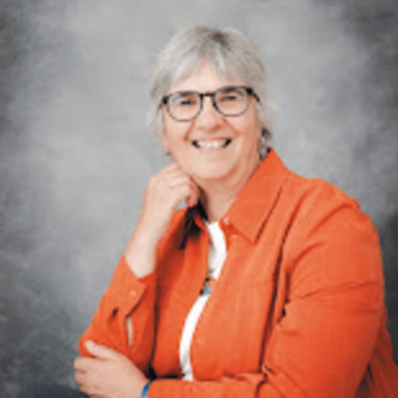 A smiling woman with gray hair and glasses, wearing an orange shirt, is posed against a gray, mottled background.