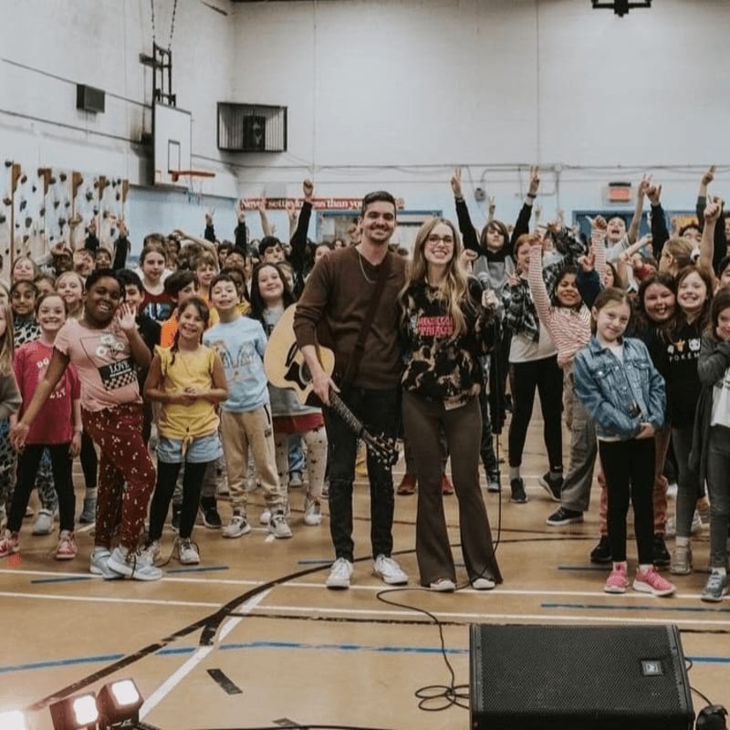A man and woman with a guitar stand in the foreground of a gymnasium filled with cheering children.