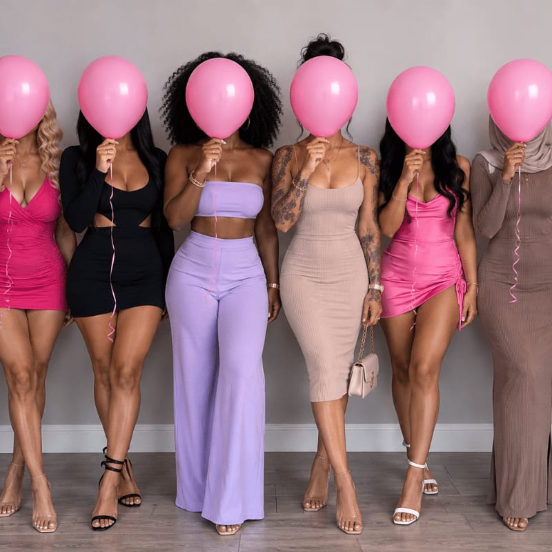 Six women in stylish outfits stand in front of a plain gray wall, each holding a pink balloon that obscures their faces.