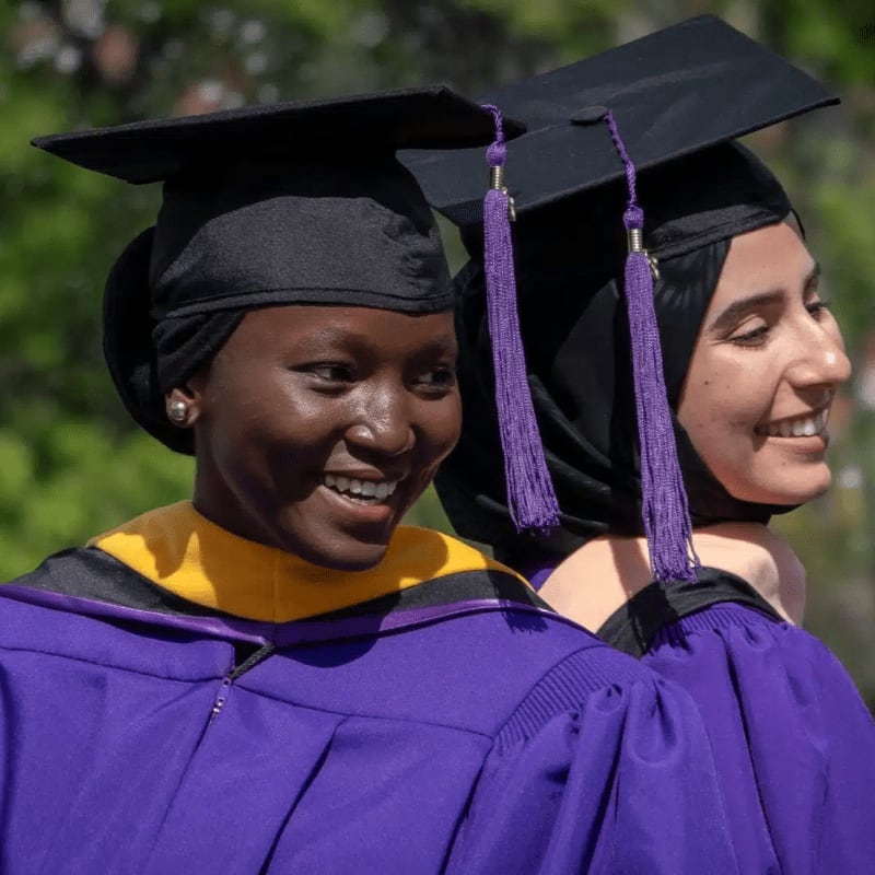 Two smiling graduates in purple gowns and black caps with purple tassels are in the foreground, with a blurred green background of trees.