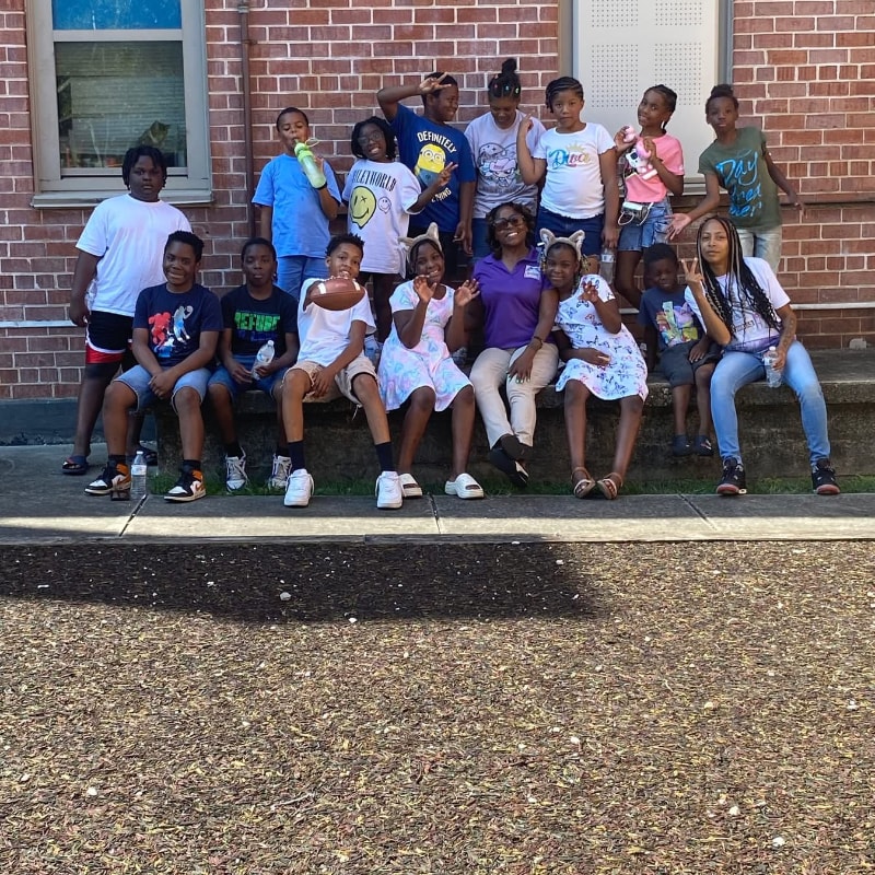 A group of children and an adult pose for a photo in front of a brick building, with a textured ground in the foreground.