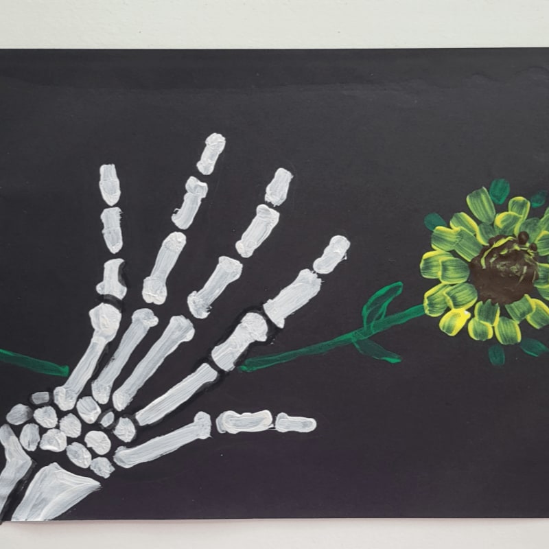 A white skeleton hand reaches towards a yellow and green flower against a black background.