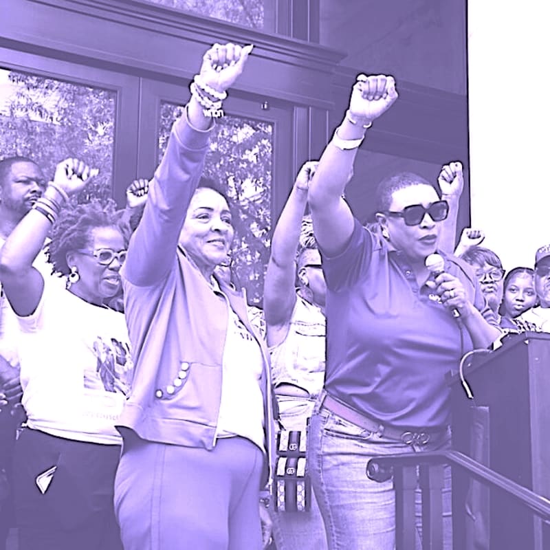 Two women raise their fists in solidarity at a rally, with a crowd of supporters visible behind them.