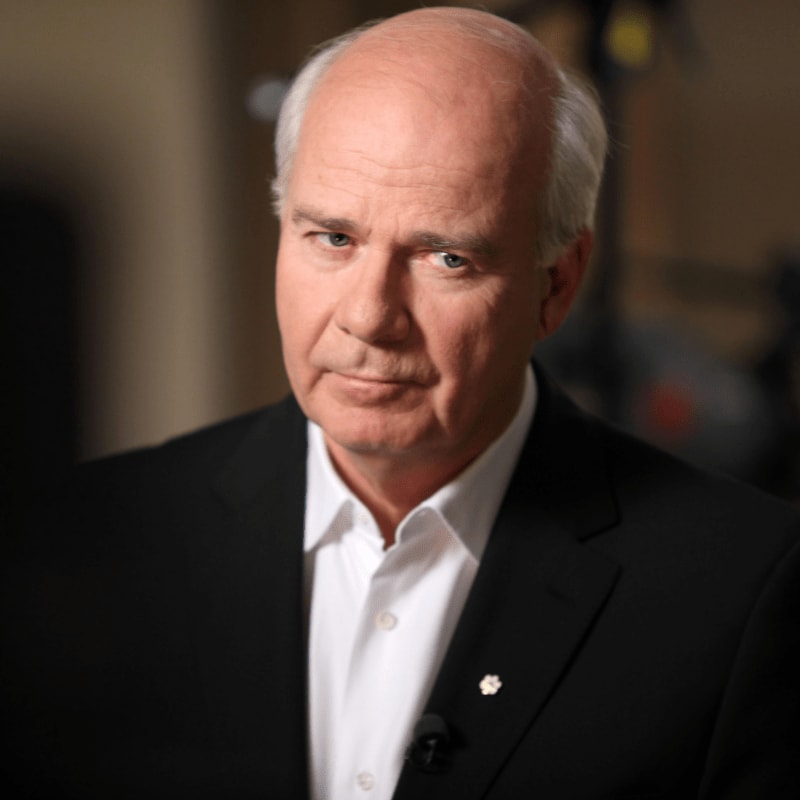 A man in a suit with a serious expression is in the foreground, with a blurred background that appears to be an indoor setting.