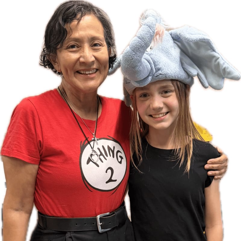 A smiling woman in a red "Thing 2" shirt stands with a young girl wearing an elephant hat, both against a plain white background.