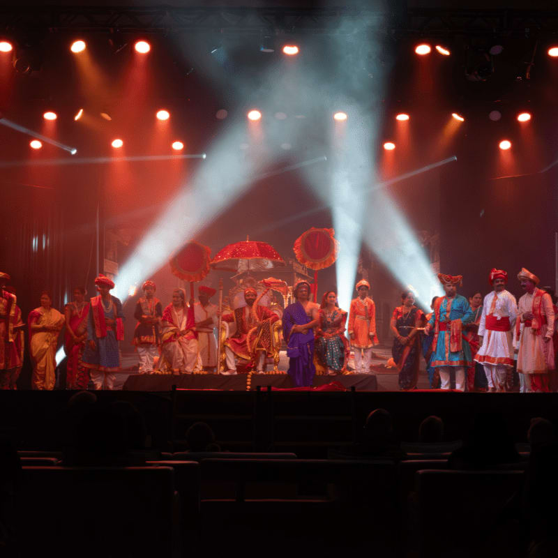 A group of performers in traditional attire stand on a stage with spotlights illuminating them, while an audience is silhouetted in the dark foreground.