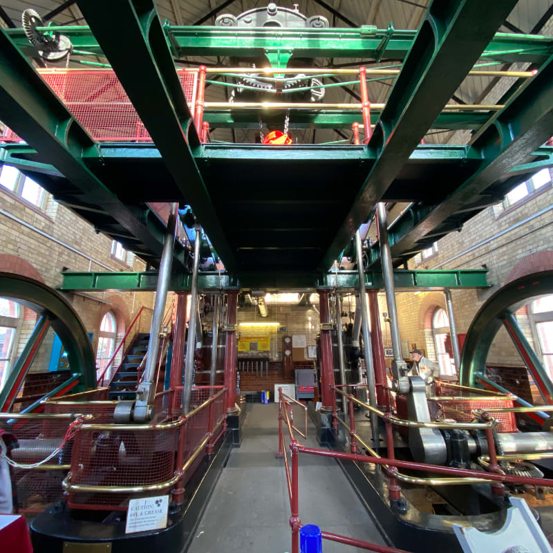 In the foreground, two large, red-railed industrial engines stand side-by-side, while the background reveals a brick building with arched windows and a complex overhead structure.