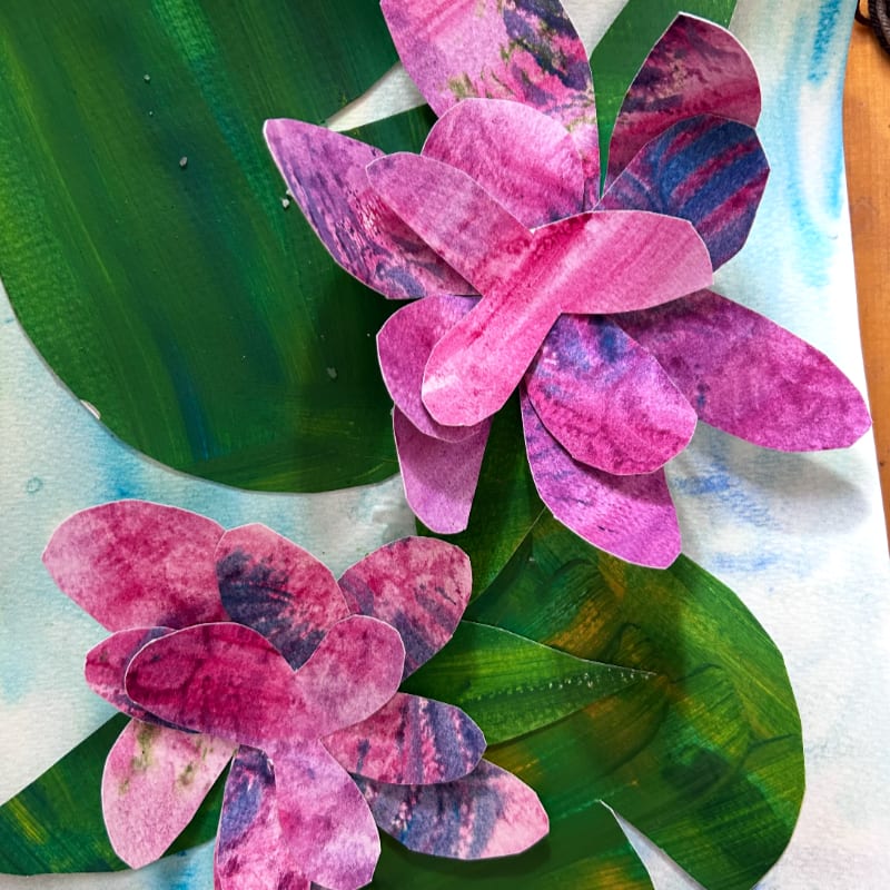 Two vibrant pink and purple paper flowers with textured petals bloom above large green leaves against a soft blue and white watercolor background.