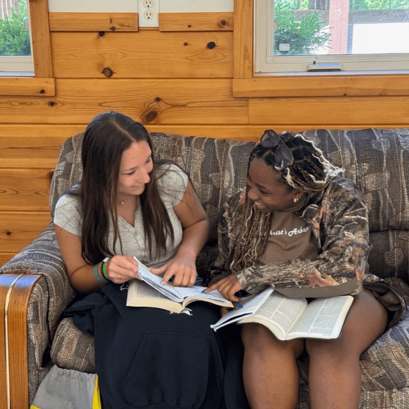 Two young women are studying books on a couch in a room with wood-paneled walls and windows.