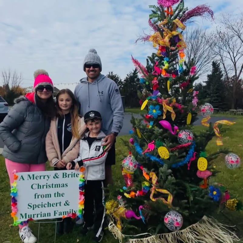 A family of four poses in front of a brightly decorated Christmas tree with a festive sign in the foreground, set against a backdrop of trees and a cloudy sky.