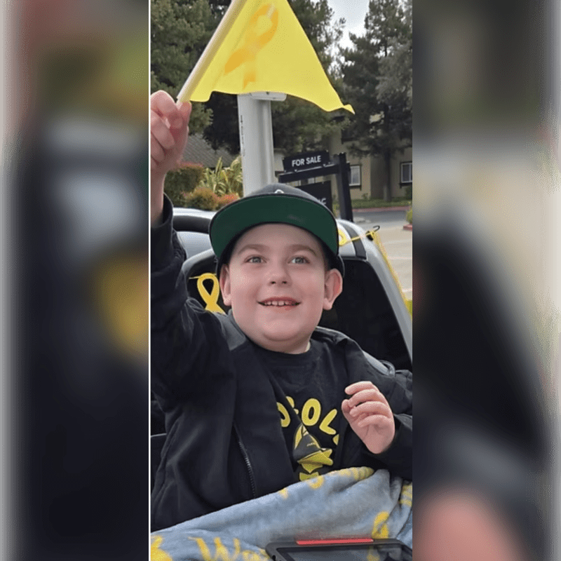 A young boy in a wheelchair smiles and waves a yellow ribbon flag, with a "for sale" sign visible on a house in the blurred background.