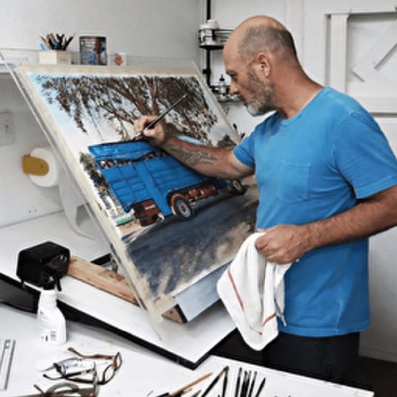 A bald man in a blue t-shirt paints a blue truck on an easel, with art supplies scattered on the table in the foreground and shelves of art materials in the background.