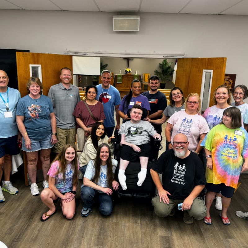 A diverse group of people, including a person in a wheelchair, pose for a photo in front of a church's wooden doors, with a cross visible in the background.