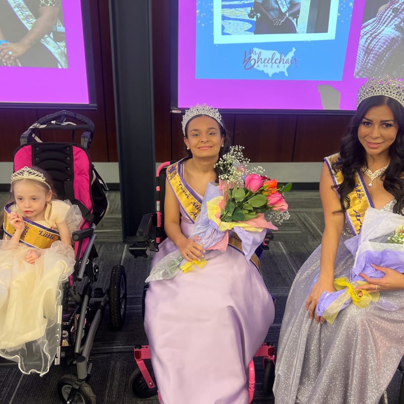 Three queens in wheelchairs, two wearing crowns and sashes, are posed in front of a projection screen displaying event information.