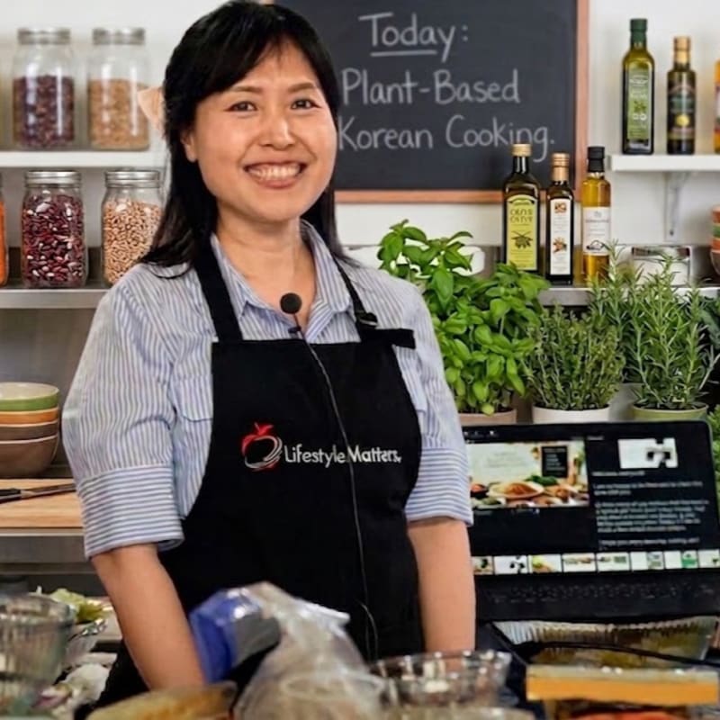 A smiling woman in an apron stands in a kitchen with shelves of ingredients and a laptop displaying a recipe in the background.