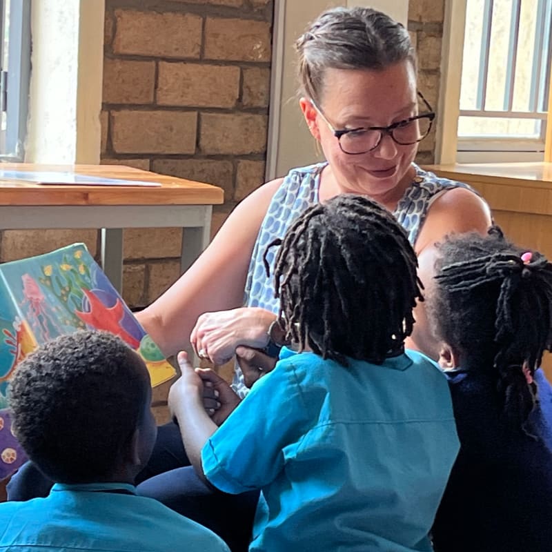 A woman reads a colorful book to three young children in a classroom with brick walls.