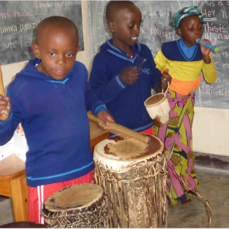 Three children play drums and sing in front of a chalkboard with writing.