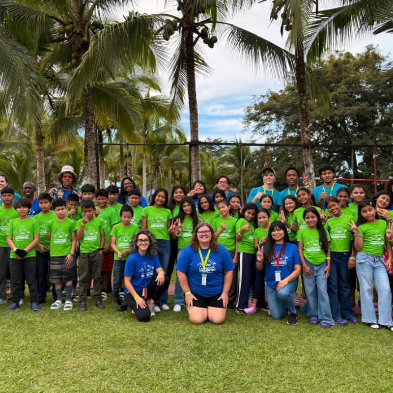 A group of children in bright green shirts pose for a photo on a grassy lawn, with palm trees and a fence in the background.