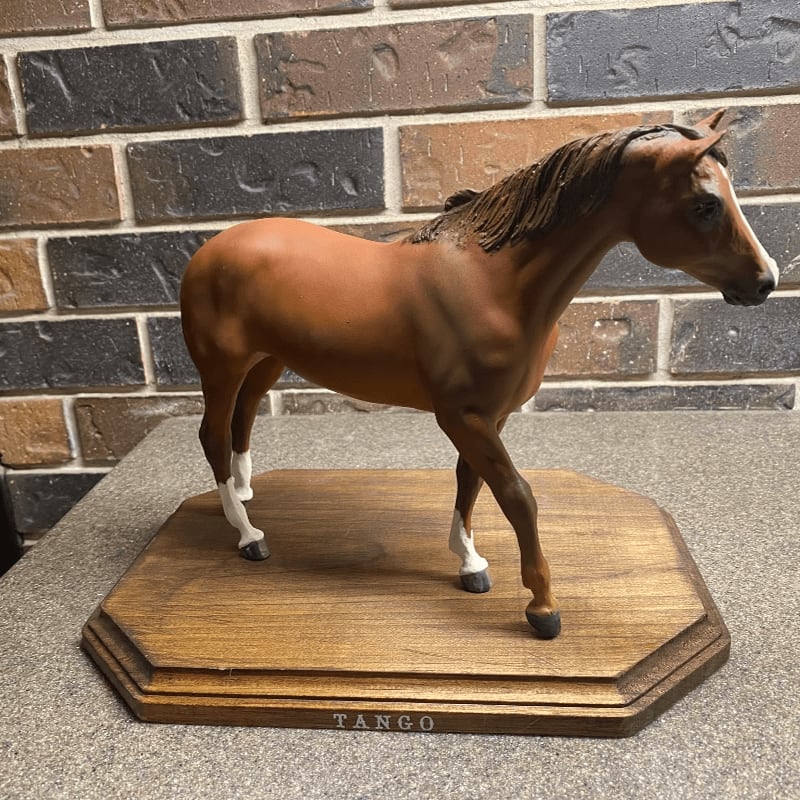 A brown horse figurine stands on a wooden base in front of a brick wall.
