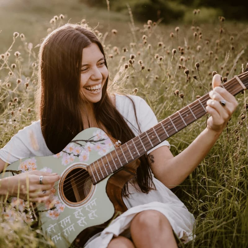 A smiling woman with long dark hair plays a decorated acoustic guitar while sitting in a field of dry grass and wildflowers.