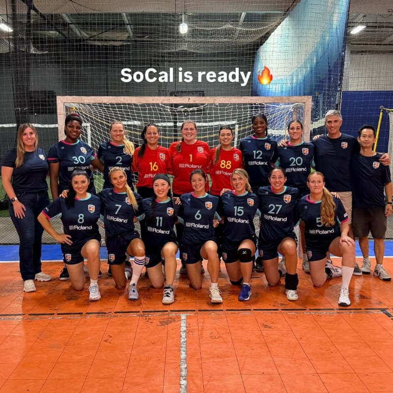 A team of women in matching uniforms poses for a photo on an orange court in front of a net, with a coach and another man standing behind them in an indoor sports facility.