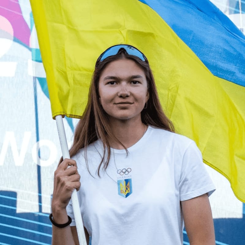 A young woman in a white t-shirt with an Olympic logo stands holding a Ukrainian flag, with a blurred blue and white background.