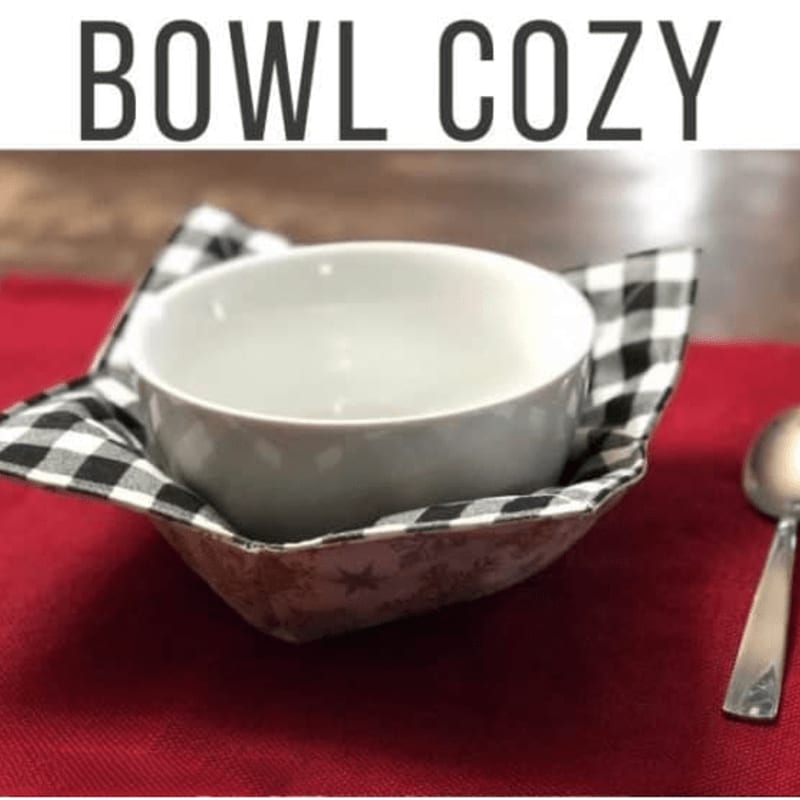 A white bowl sits in a black and white checkered bowl cozy on a red placemat, with a spoon to the right.