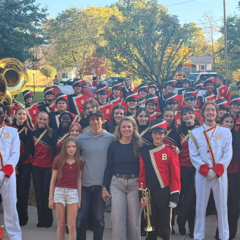 A group of band members in uniform stand behind a man and two children in the foreground, with trees and houses in the background.