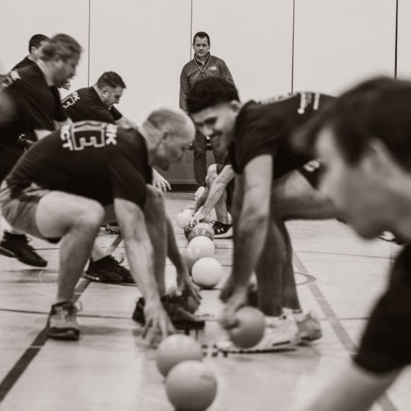 In the foreground, players crouch low to grab dodgeballs on a gymnasium floor, while in the background, a man stands watching.
