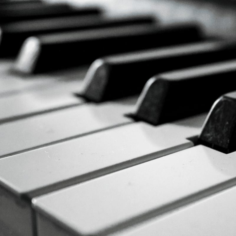 A close-up, black and white shot of piano keys fills the frame, with the foreground showing the white keys in sharp focus and the black keys receding into a blurred background.