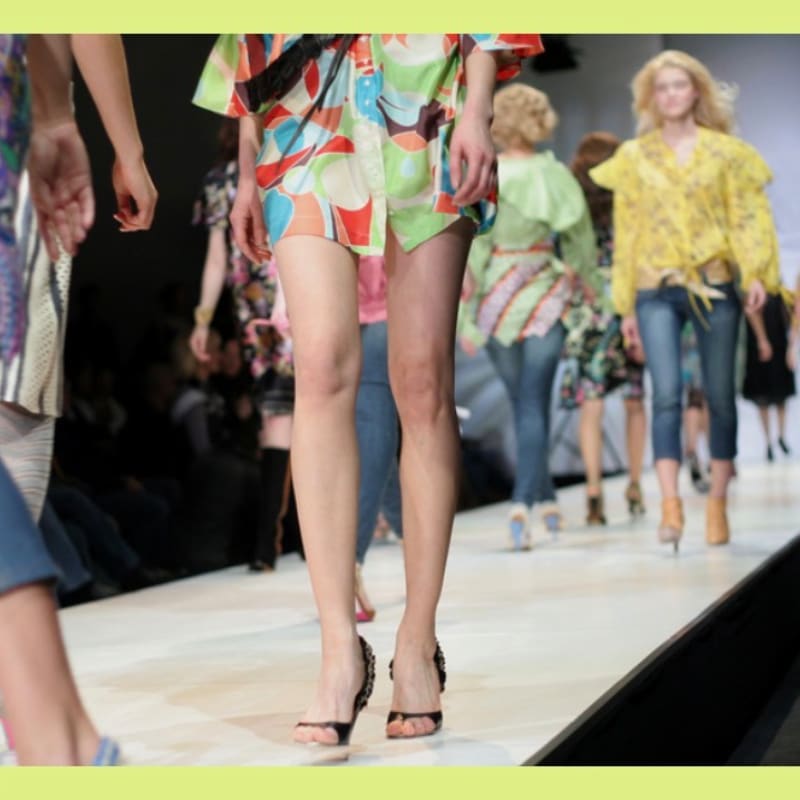 Models with colorful outfits walk down a runway with an audience blurred in the background.