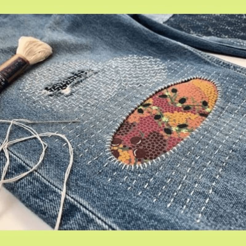 A needle and thread lie next to a denim patch being mended with visible stitching, while a colorful fabric patch is sewn onto the jeans in the background.