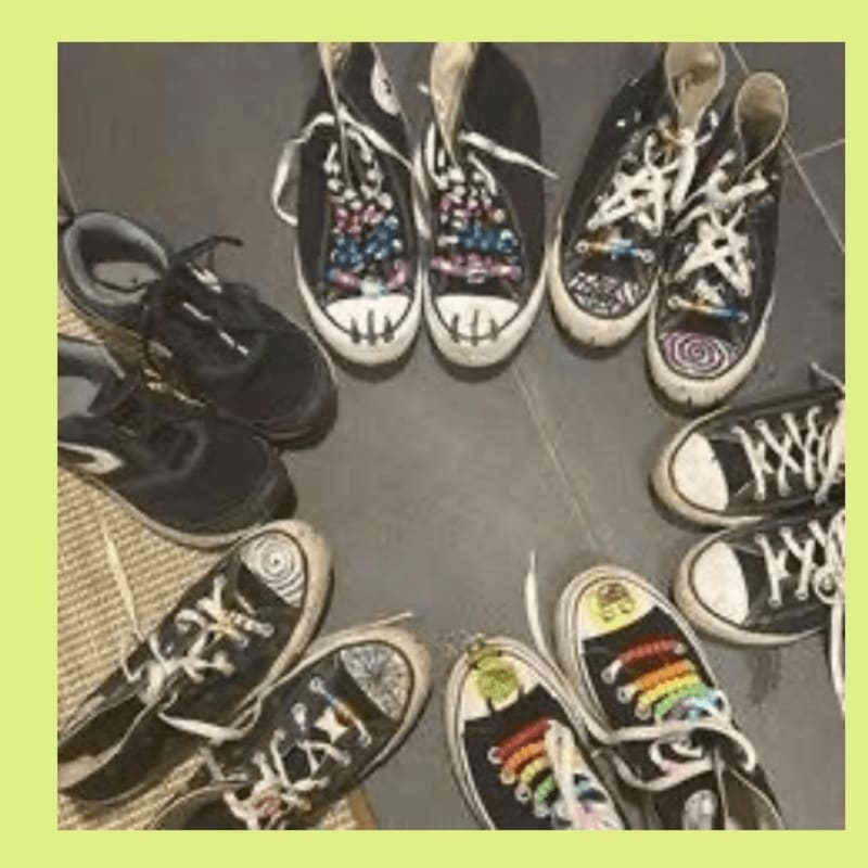 Several pairs of black sneakers, some with colorful laces and designs, are arranged in a circle on a dark floor, with a woven mat visible in the lower left corner.