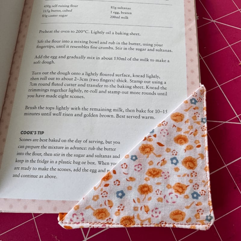 A floral fabric corner bookmark rests on an open cookbook page with scone recipes, set against a pink cutting mat.