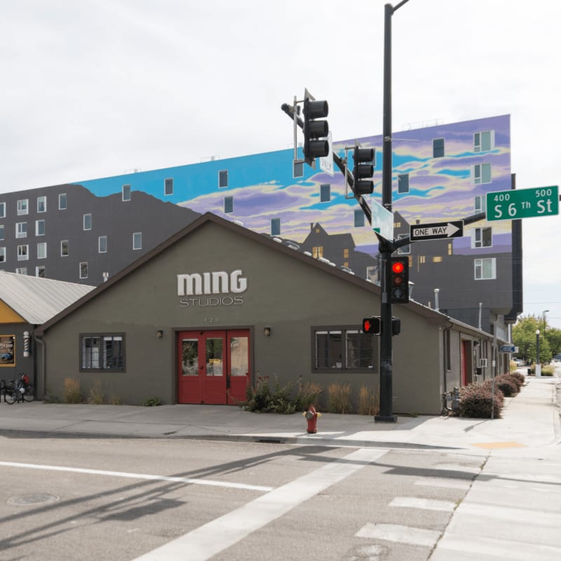 A low-rise building with "ming STUDIOS" on its facade stands in the foreground, while a tall building with a colorful mural of a sky and clouds rises in the background.