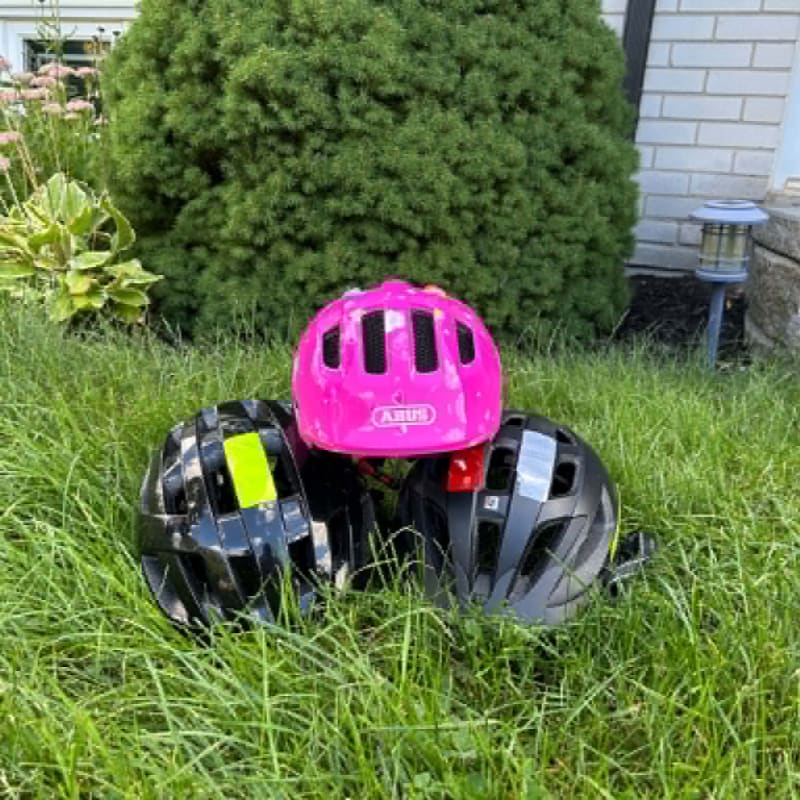 Three bicycle helmets, one pink and two dark-colored, rest on lush green grass in front of a large, manicured bush and a house.
